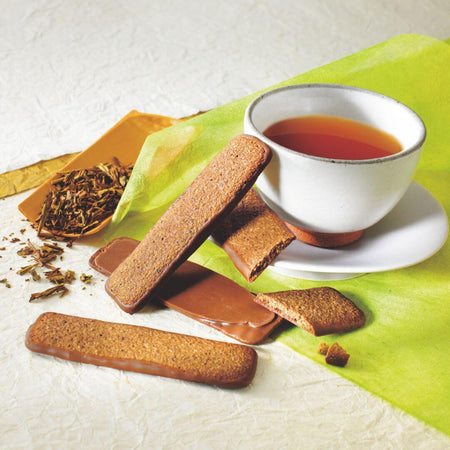 ROYCE' Chocolate - ROYCE' Kyoto Baton Cookies "Hojicha" - Image shows brown, chocolate-coated, baton-shaped cookies on a white surface with green paper. Accents include a tea cup and some loose tea leaves.