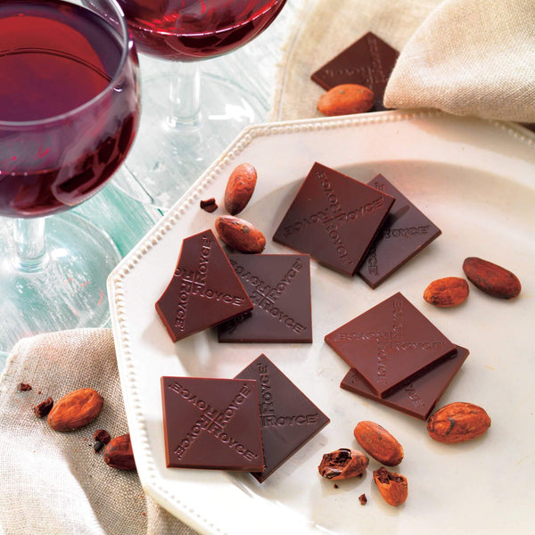 ROYCE' Chocolate - ROYCE' Origin Chocolate "Bitter" - Image shows brown chocolate squares with the words "ROYCE'" engraved. Accents include a white hexagon-shaped plate, loose cacao beans, a tablecloth in light yellow, and two glasses filled with red wine. Background and surface is wood painted in light blue.