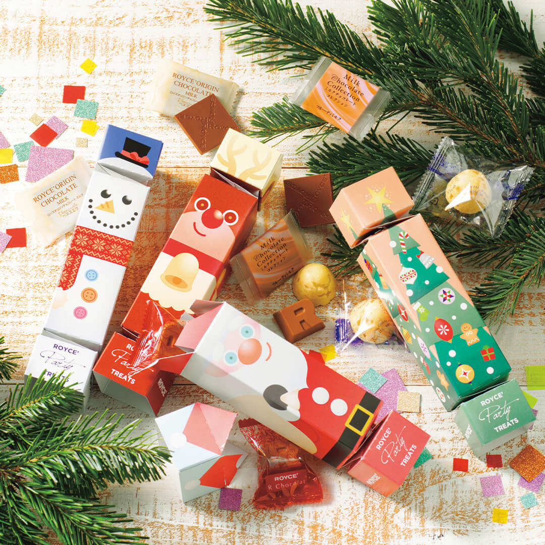 ROYCÉ Party Treats: Christmas-themed cracker gift boxes with assorted Royce chocolates, including milk and strawberry pieces, among festive pine sprigs and confetti.