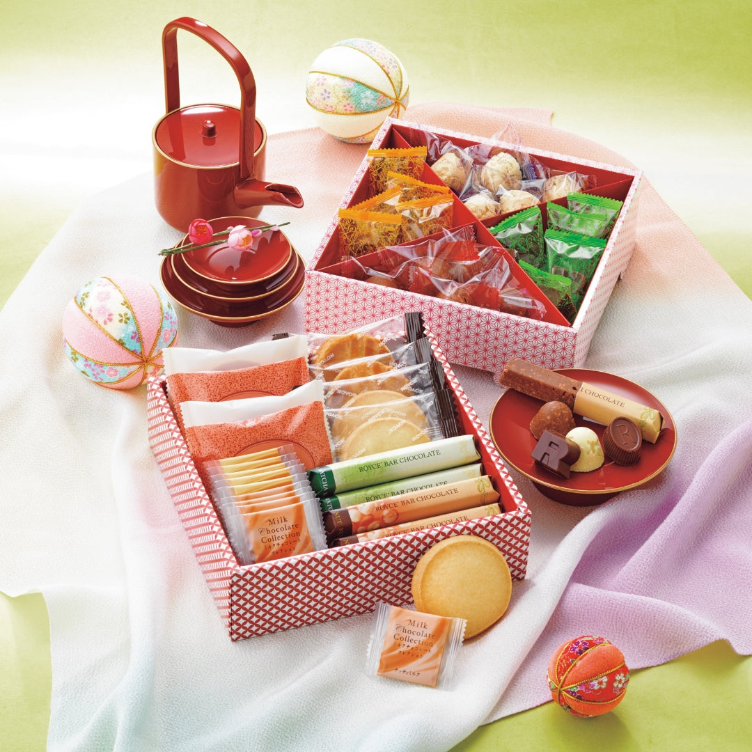 ROYCE' Sweets Osechi - Image shows open printed boxes with individually-wrapped confections inside. Accents include red plates and a red tea pot. Also seen are decorative balls and loose confections.