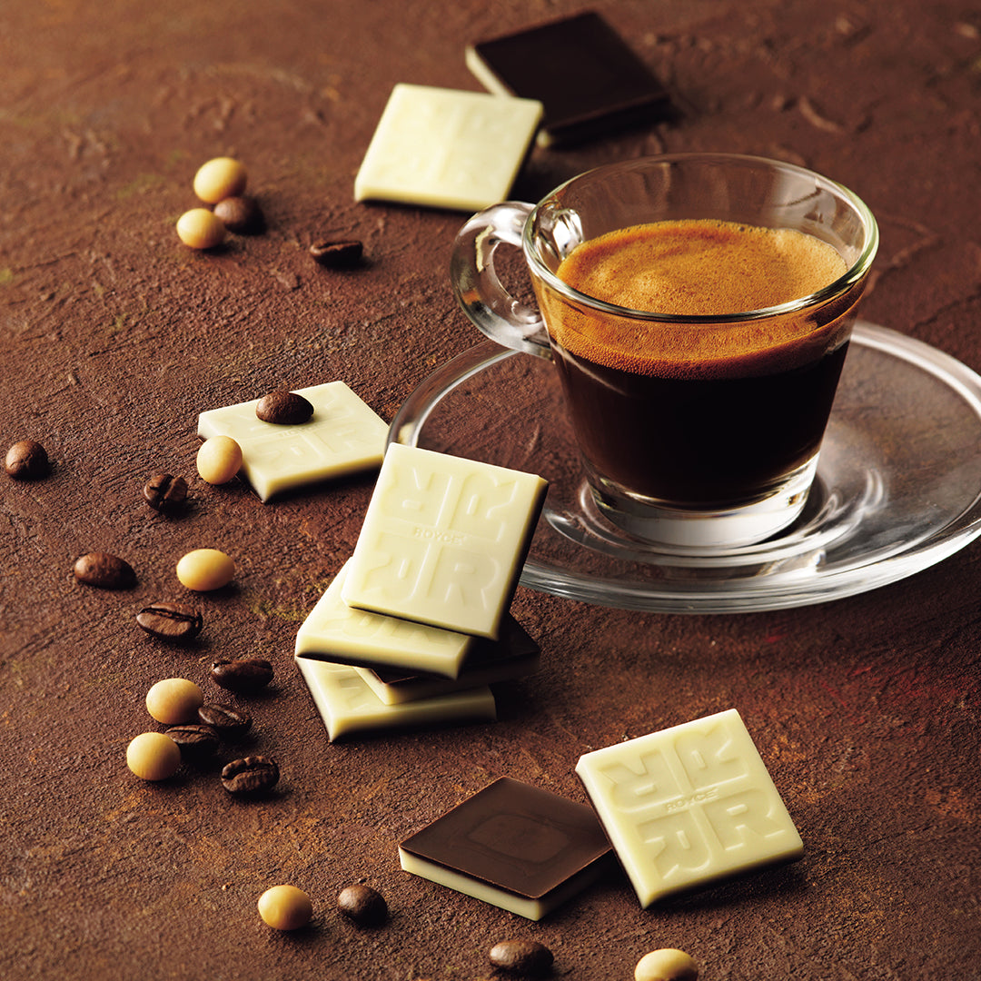 Soy Espresso Chocolate - Image shows two-layered chocolate squares on a brown surface with accents of loose coffee and soy beans. Also seen is a cup of espresso.
