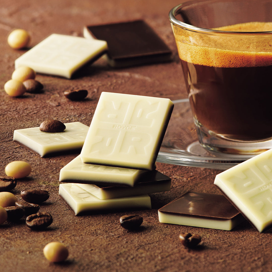 Soy Espresso Chocolate - Image shows two-layered chocolate squares on a brown surface with accents of loose coffee and soy beans. Also seen is a cup of espresso.