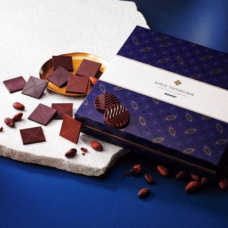 ROYCE' Chocolate - ROYCE' Tasting Box - Image shows a blue box with cacao prints and a white center. Text in center says ROYCE' Tasting Box ROYCE'. Accents include loose chocolates, a white platform, and a small gold plate. Background is in blue.