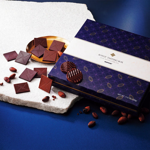 ROYCE’ Tasting Box with embossed chocolates, wavy discs, and cocoa beans, showcased in front of a decorative box with geometric accents and brand logo.