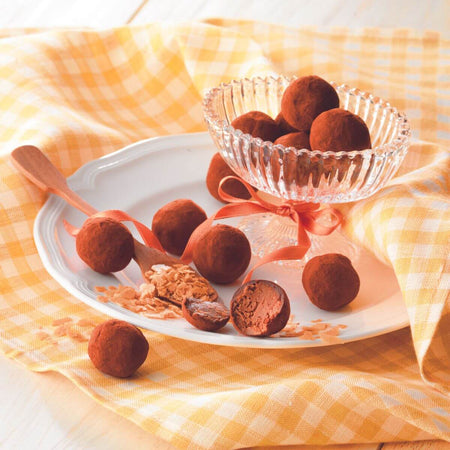 ROYCE' Chocolate - ROYCE’ Feuillantine Truffe “Gianduja” - Image shows brown chocolate truffles on a plate and glass. Accents include a wooden spoon and loose baked flakes together with a tablecloth in yellow and white.