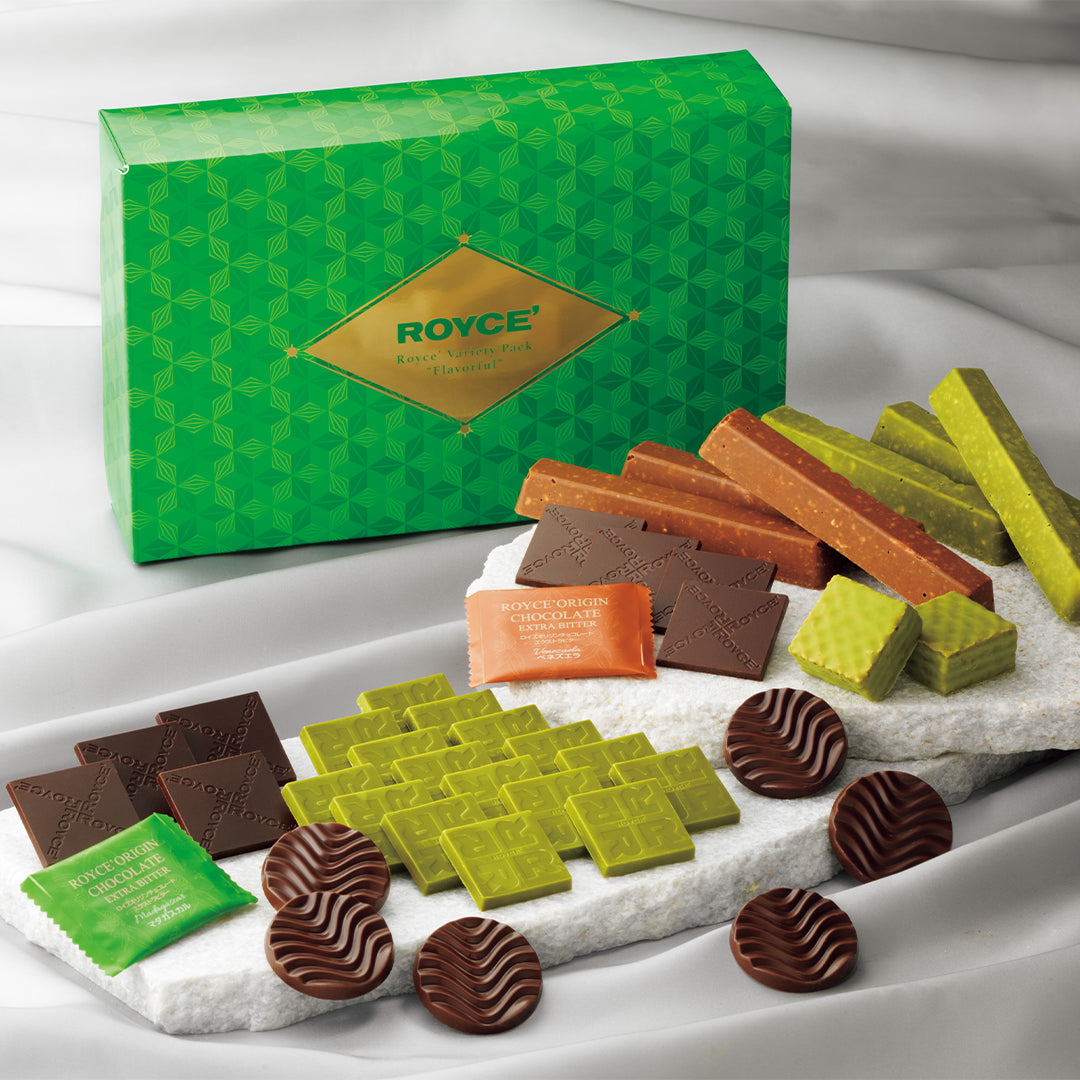 ROYCE' Variety Pack "Flavorful" - A boxed assortment of chocolates and sweets displayed on a white draped fabric background. The green ROYCE' gift box stands behind the confectionery items, which include dark and milk chocolate pieces, green and orange wrapped pieces, and striped chocolate candies arranged on a white stone platter.