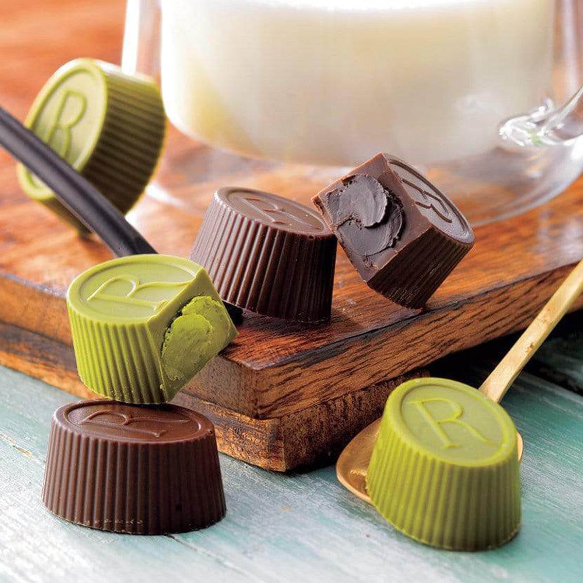 ROYCE' Chocolate - Tea Chocolat "Hojicha & Matcha" - Image shows brown and green chocolate cups engraved with the letter R. Accents include brown wooden trays and spoons. Background features a glass of milk and a wooden surface in color blue.