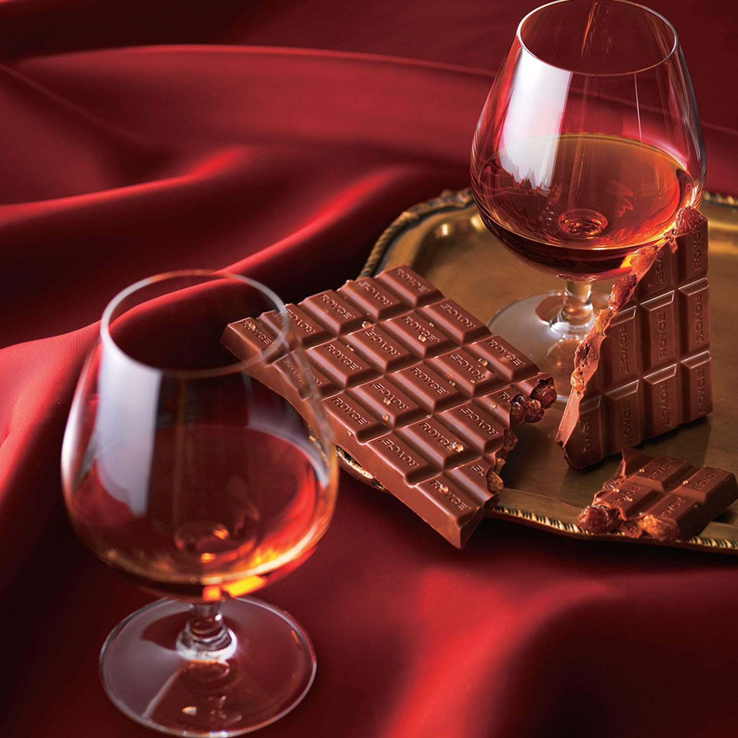 ROYCE' Chocolate - Chocolate Bar "Cognac & Raisin" - Image shows two glasses filled with amber-hued liquid and two brown chocolate bars with the word ROYCE' engraved multiple times on each. Accents include a gold tray and a silky, rippled red tablecloth for background.