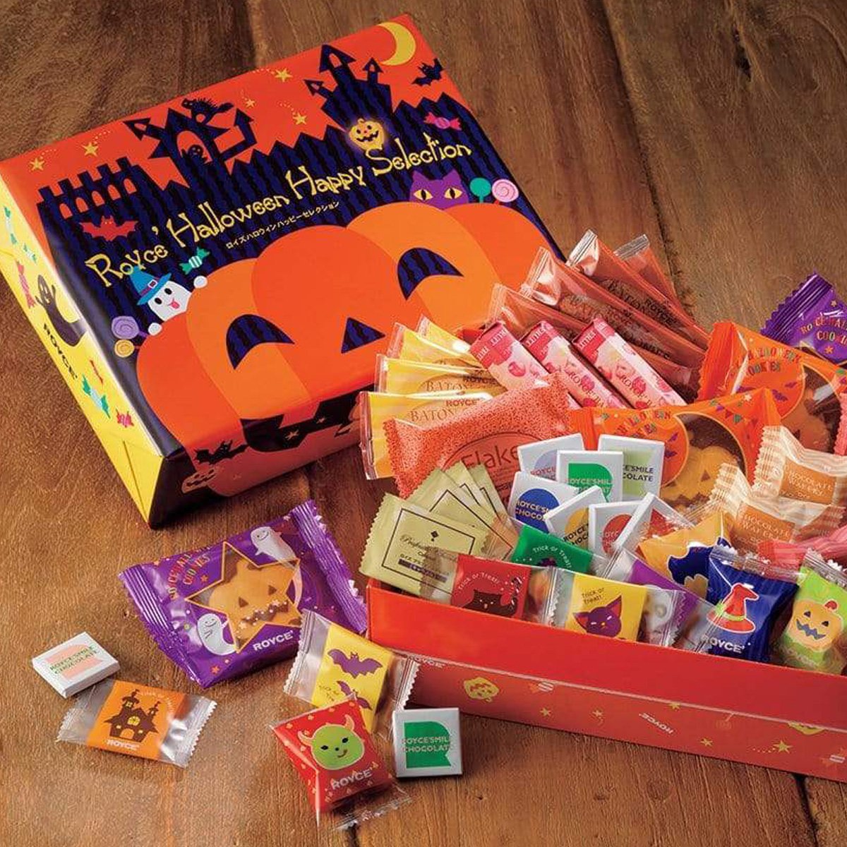 Image shows an orange box on the left with illustrations of ghosts, animals, stars, and pumpkins. Text says ROYCE' Halloween Happy Selection. Box on the right shows individually-wrapped chocolates in various prints and colors. Background is in brown wood.