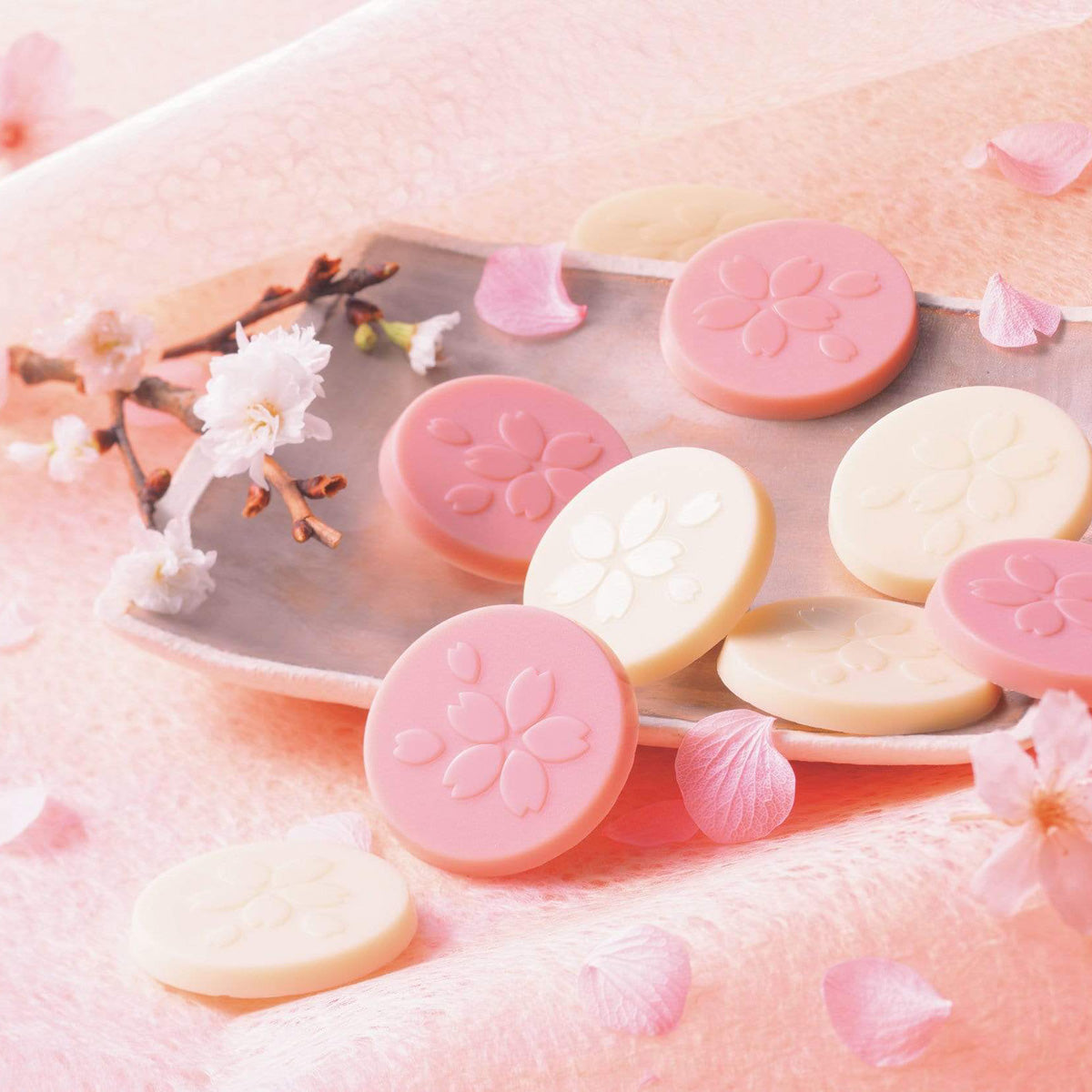 ROYCE' Chocolate - Sakuraberry & Sakurawhite Chocolate - Image shows pink and white chocolate discs with floral engravings. Accents include a gray plate, white flowers, pink petals, and brown twigs. Background is in pink color.