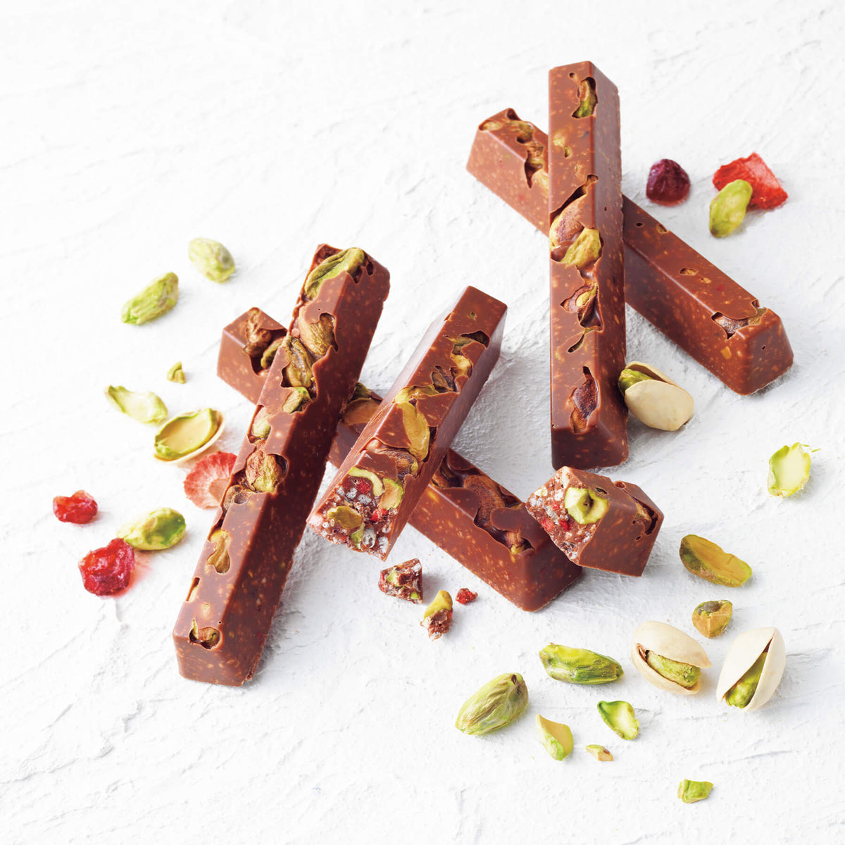 ROYCE' Chocolate - ROYCE' Bar Chocolate "Pistachio" - Image shows finger-shaped brown chocolate bars with pistachio nuts and berries inside. Accents include loose pistachio nuts and dried berries. Textured background is in white.