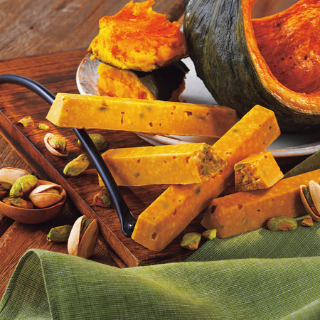 Image shows yellow finger-shaped chocolate bars on a brown wooden tray and table. Accents include a brown wooden scoop, loose pistachio nuts in green and light yellow, a green tablecloth, and a white plate with pumpkin slices in the colors of green and yellow-orange. Background is in brown color.