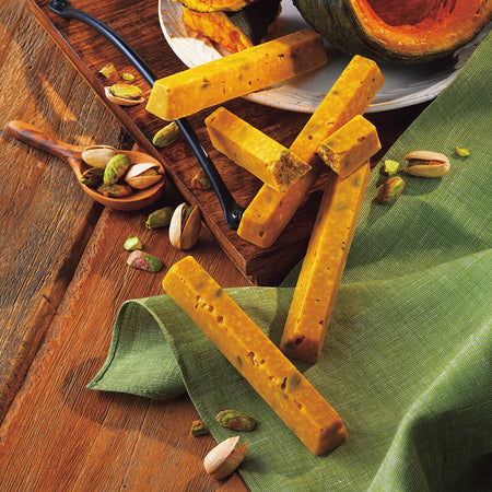 Image shows yellow finger-shaped chocolate bars on a brown wooden tray and table. Accents include a brown wooden spoon, loose pistachio nuts in green and light yellow, a green tablecloth, and a white plate with pumpkin slices in the colors of green and yellow-orange.