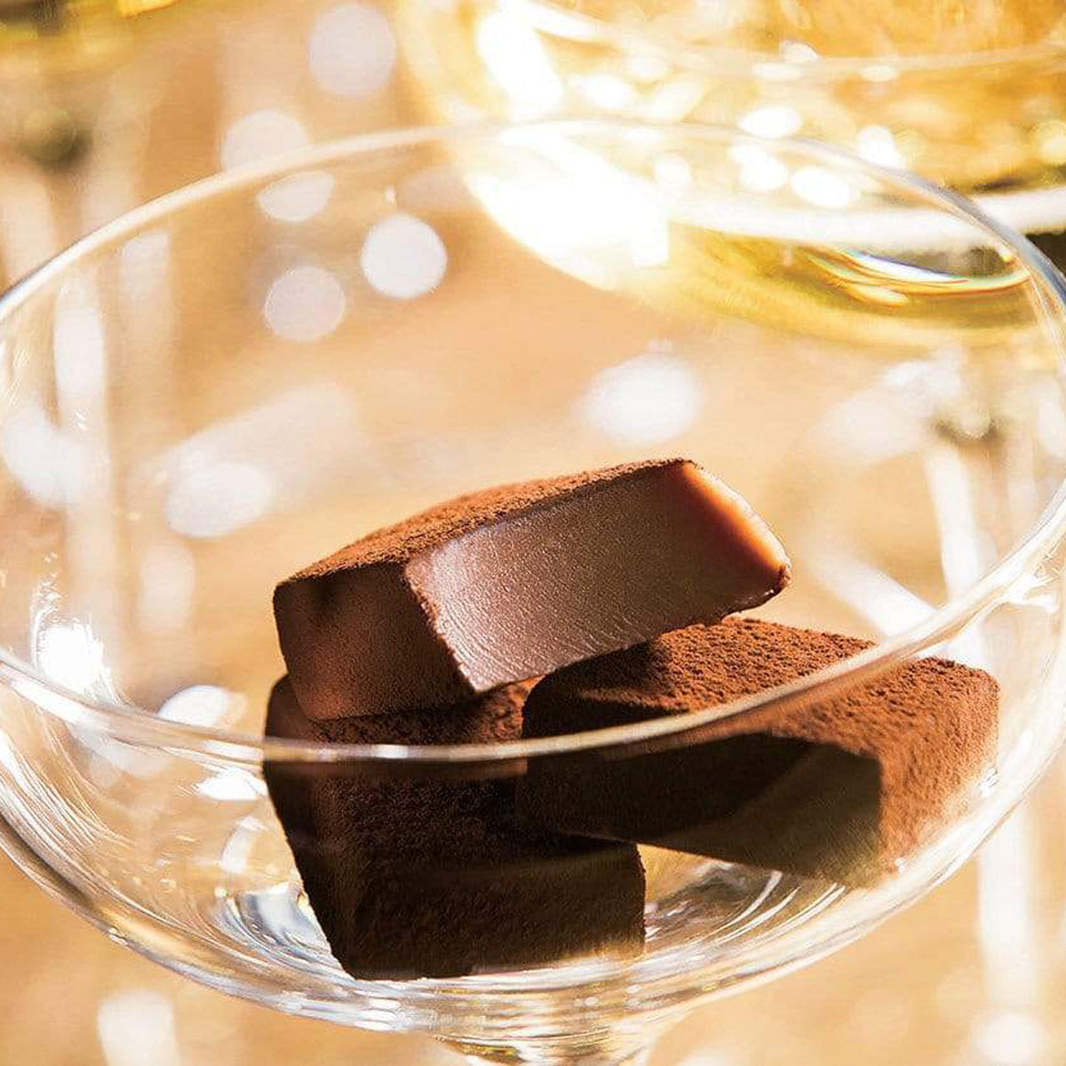 ROYCE' Chocolate - Nama Chocolate "Champagne Pierre Mignon" - Image shows brown blocks of chocolates in a clear champagne glass. Background is yellow and there is an accent of a blurry glass filled with yellow champagne.