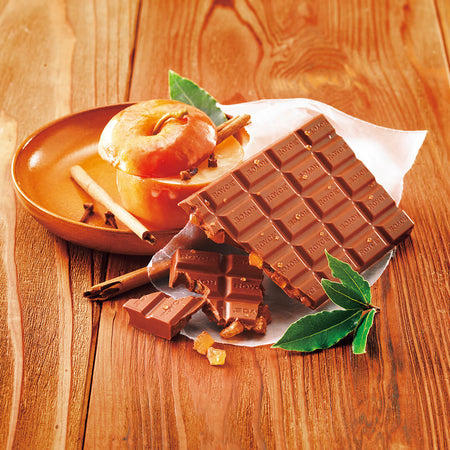 ROYCE' Chocolate - Chocolate Bar "Baked Apple" - Image shows a brown chocolate bar on a crumpled white paper. Accents include a brown wooden bowl with a caramelized apple and some cinnamon rolls. Also seen are green leaves. Background is light brown.