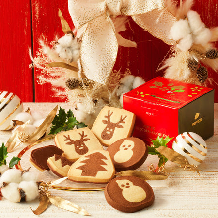 Christmas Cookies 6 Pcs - Image shows a plate filled with cookies in brown and yellow in different shapes with illustrations of reindeer, a tree, and snowmen. Red and green box on right has gold prints of leaves. Text says ROYCE' Christmas Cookies ROYCE'. Accents include striped decoration balls in gold and white, golden ribbons, leaves, stems, and acorns. There are also white cotton balls for a snow-like effect. Background is in red and surface is in white.
