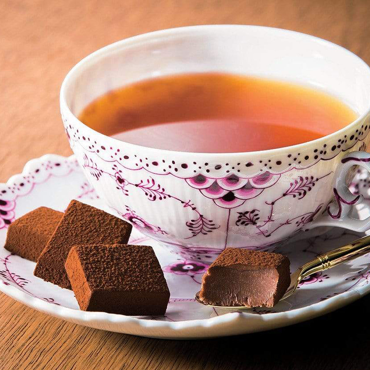 ROYCE' Chocolate - Nama Chocolate "Darjeeling" - Image shows brown blocks of chocolate on a a white plate with pink floral prints. Accents include a white tea cup with pink floral prints filled with amber-hued tea and a brown wooden background.