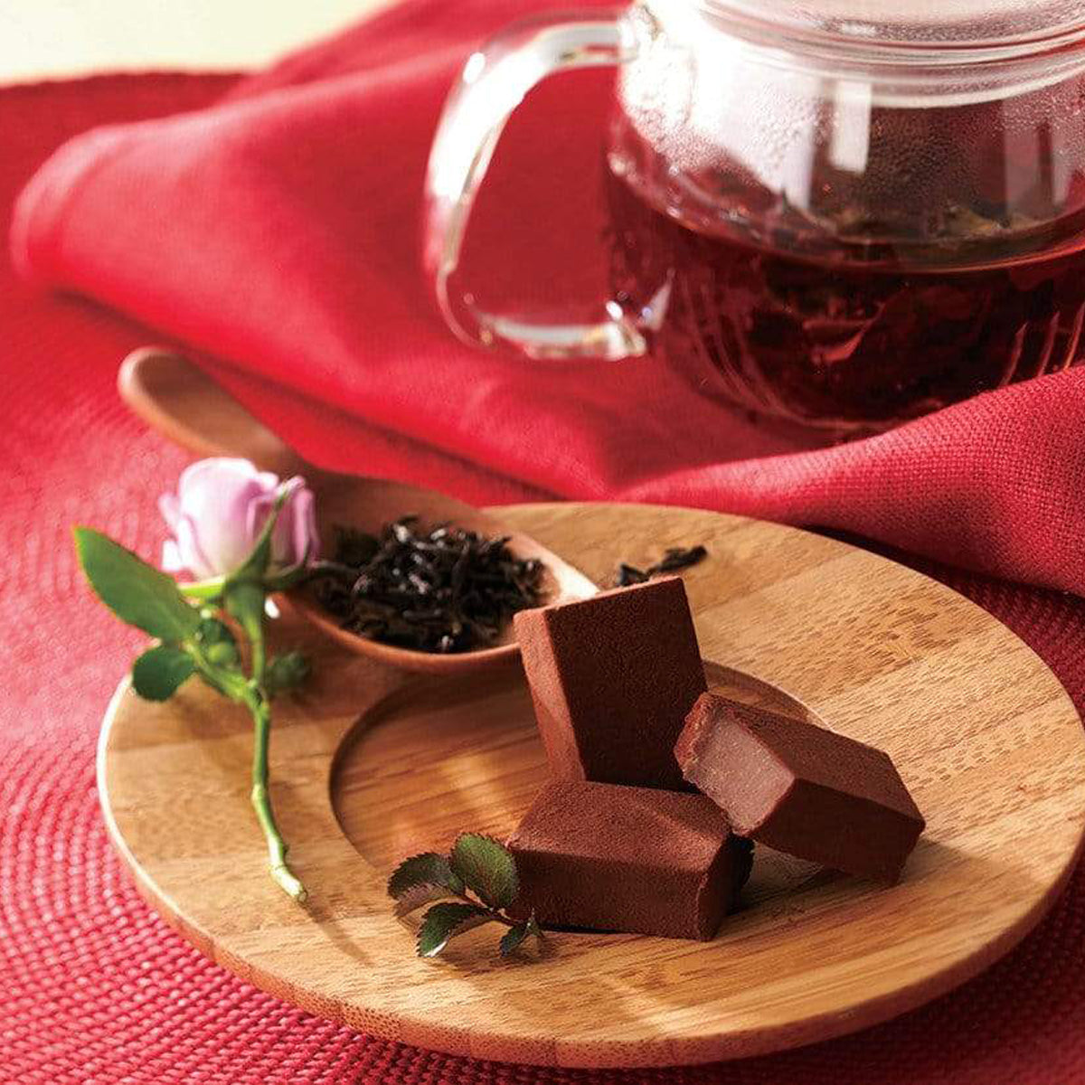 ROYCE' Chocolate - Nama Chocolate "Darjeeling" - Image shows brown blocks of chocolate on a brown wooden plate. Accents include a pink rose with green stem, a wooden spoon with tea shavings, a red cloth background, and a clear teapot filled with amber-hued tea.