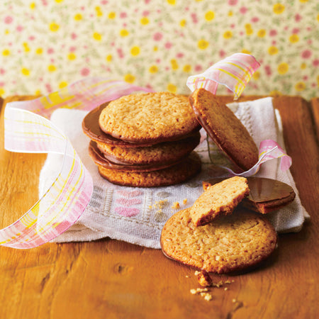 ROYCE' Chocolate - Graham Chocolate Cookie - Image shows chocolate-coated cookies on a wooden surface with a plaid ribbon and embroidered towel. Background has blurry floral prints.