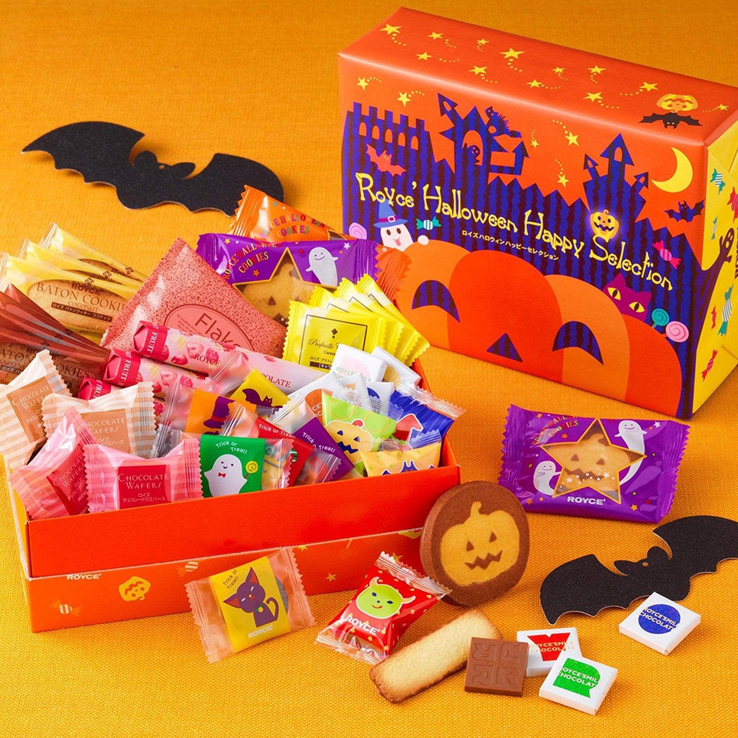 Image shows an orange box on the right with illustrations of ghosts, animals, stars, and pumpkins. Text says ROYCE' Halloween Happy Selection. Box on the left shows individually-wrapped chocolates in various prints and colors. Accents include bat-shaped black paper cutouts and some cookies and chocolates.