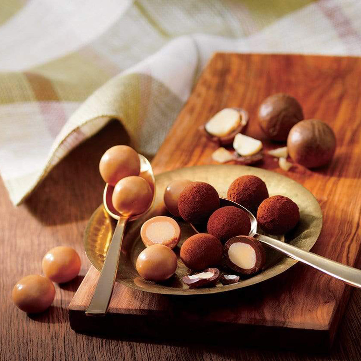 ROYCE' Chocolate - Macadamia Chocolate - Image shows brown chocolate-coated macadamia nuts on a copper plate with bronze spoons. Background accents include macadamia nuts, a brown wooden chopping board and a white cloth with red and brown stripes.