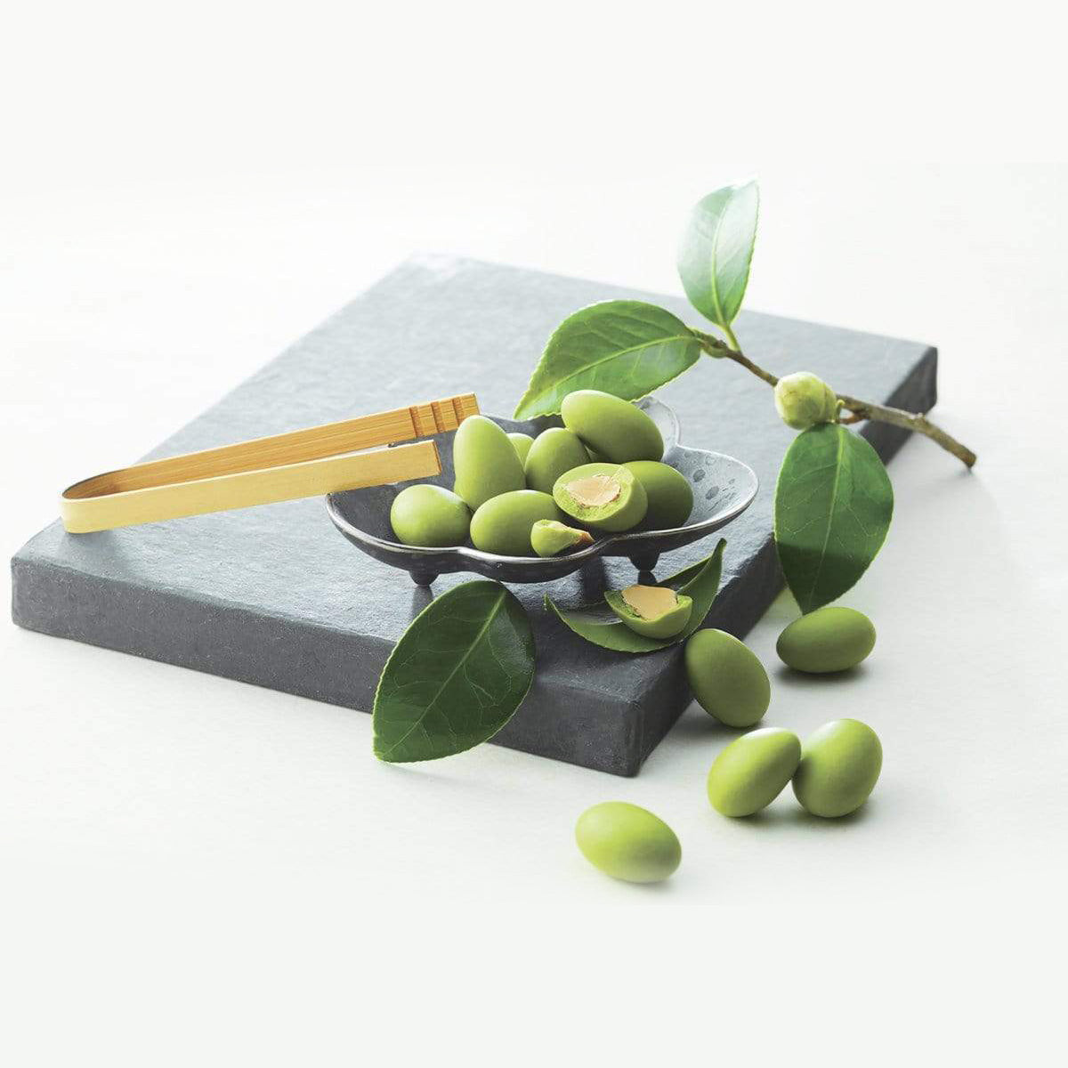 ROYCE' Chocolate - Matcha Almond Chocolate - Image shows round green chocolate-coated almonds on a gray plate and gray stone platform. Accents include a yellow wooden tong and some green leaves and stems. Background is in white.