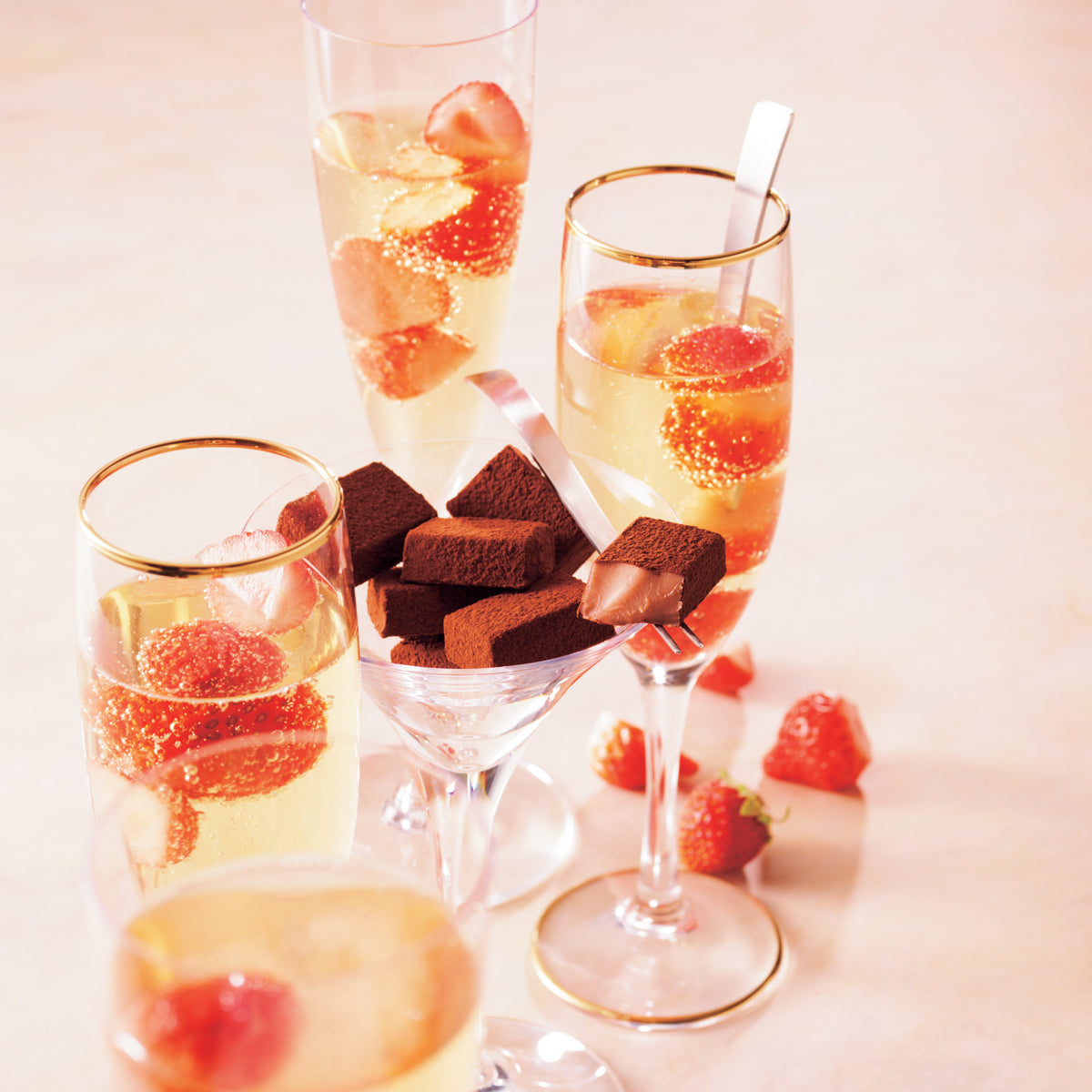 Image shows chocolate blocks on a clear cocktail glass with a light pink ribbon. Accents include champagne glasses filled with champagne and red strawberries. Other accents include red strawberries on the ground and silver cutlery used to hold a chocolate block and an extra inside a champagne glass. Background is in flesh color.