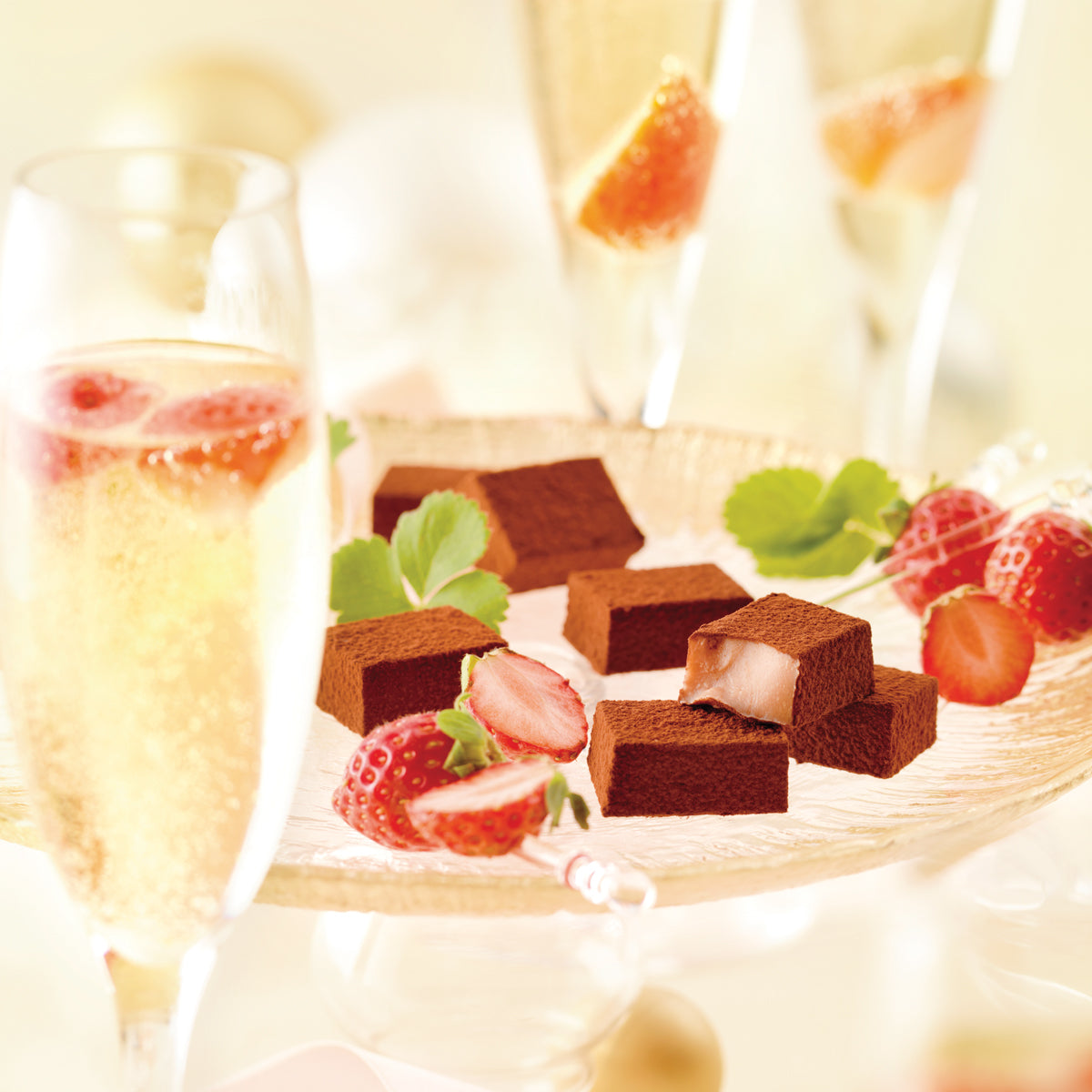 Image shows brown chocolate blocks on a clear plate with red strawberries and green leaves. Accents include champagne glasses filled with champagne and red strawberries. Background is in white.