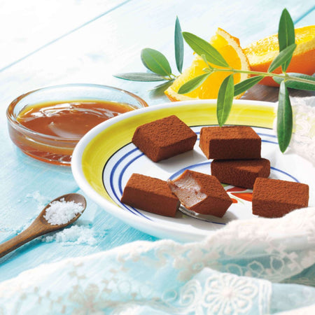 ROYCE' Chocolate - Nama Chocolate "Caramel Salé & Orange" - Image shows brown chocolate blocks on a plate colored white, yellow, and with blue lines and red floral illustrations. Accents include a wooden spoon with salt, a clear bowl with brown caramel inside, slices of oranges, and green leaves. Surface is blue wood with white lace tablecloth.