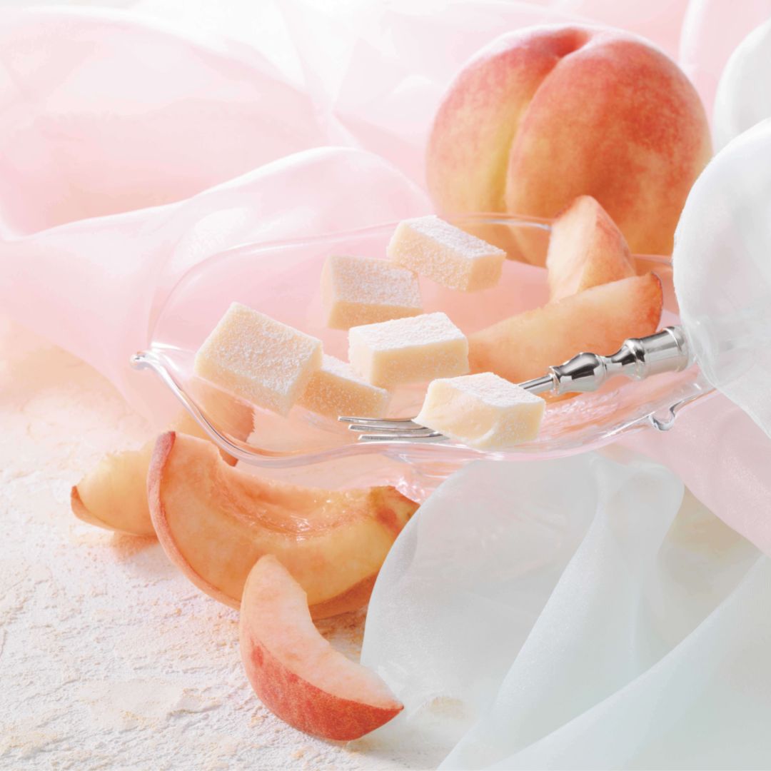 ROYCE' Chocolate - Nama Chocolate "Hakutou" - Image shows light pink chocolate blocks on a clear plate with a silver fork and peach slices. Accents include pink and white cloths and peach fruits.