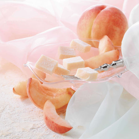 ROYCE' Chocolate - Nama Chocolate "Hakutou" - Image shows light pink chocolate blocks on a clear plate with a silver fork and peach slices. Accents include pink and white cloths and peach fruits.