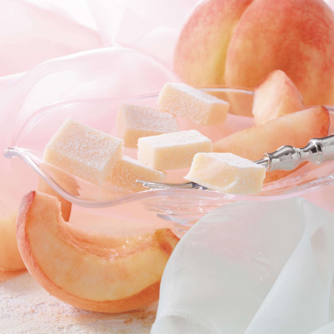 ROYCE' Chocolate - Nama Chocolate "Hakutou" - Image shows light pink chocolate blocks on a clear plate with a silver fork and peach slices. Accents include pink and white cloths and peach fruits.