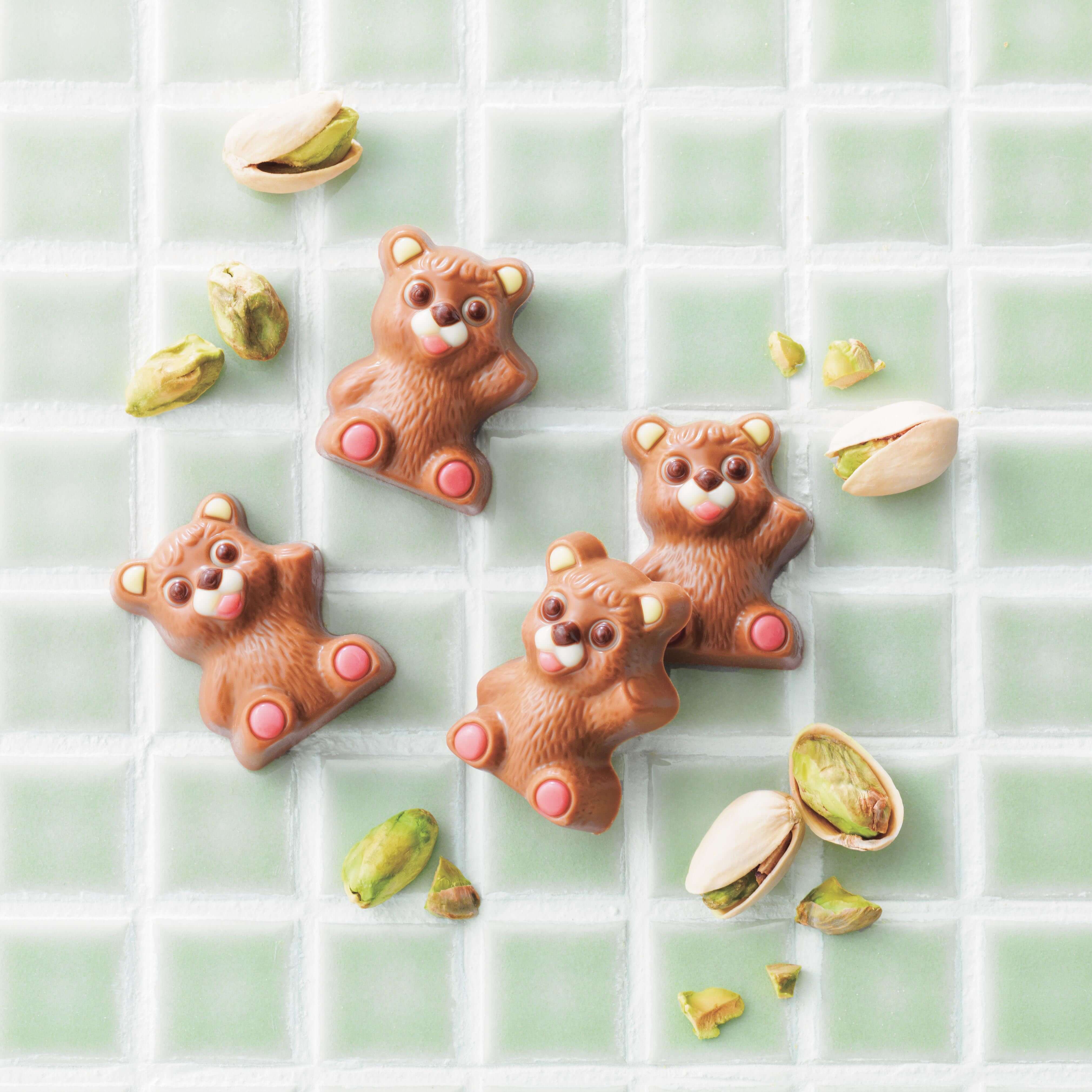 ROYCE' Chocolate - Petit Bear Chocolat "Pistachio (10 Pcs)" - Image shows brown, bear-shaped chocolates with accents of loose pistachio green nuts and beige shells. Background features green tiles with white spaces in between,