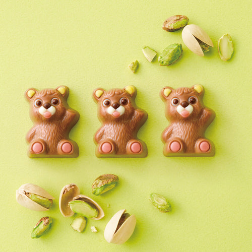 Petit Bear Chocolat Pistachio (10 Pcs) features three bear-shaped milk chocolates filled with pistachio cream, accented with raspberry-infused white chocolate, surrounded by scattered pistachios.