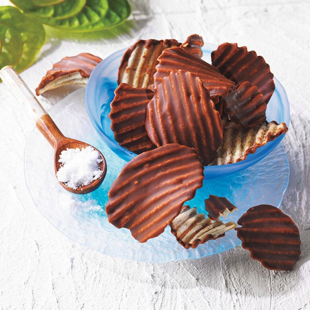 ROYCE' Chocolate - ROYCE' Ishigakijima Potatochip Chocolate "Ishigaki no Shio" - Image shows chocolate-coated potato chips on a blue bowl and clear plate. Accents include a wooden spoon with salt and green leaves. Background is a white surface.