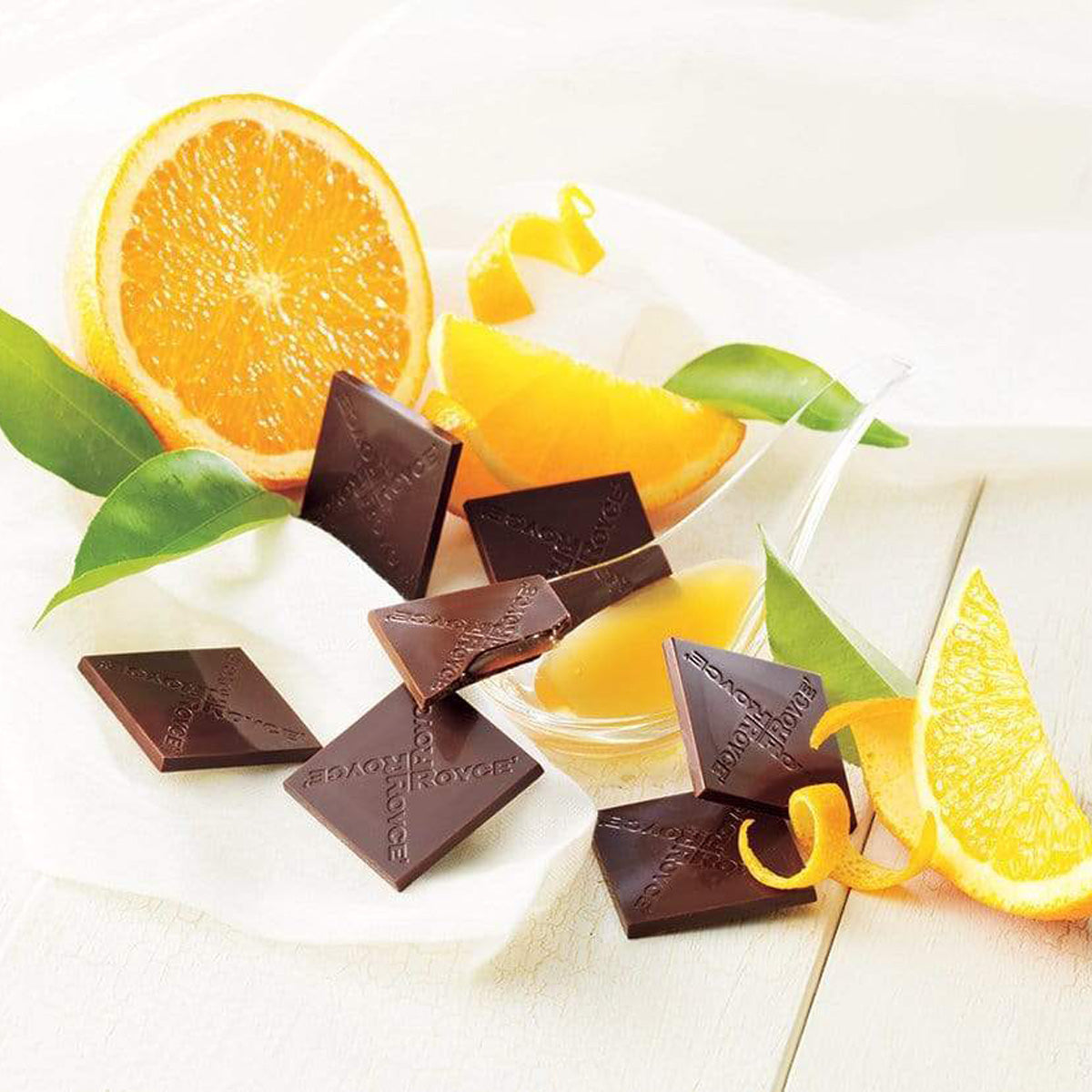 ROYCE' Chocolate - Prafeuille Chocolat "Orange" - Image shows brown chocolate squares filled with orange sauce and with the words "ROYCE'" engraved, as placed on a wooden surface in white. Accents include fruit slices of orange and orange skin peel, green leaves, a clear plastic spoon with orange sauce, and a white tablecloth. Background is in color white.