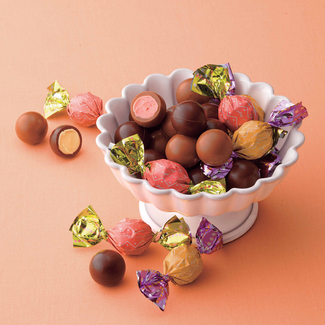ROYCE' Chocolate - ROYCE' Petit Ball Chocolat "Strawberry & Lemon" - Image shows a variety of round-shaped chocolates in different colors both wrapped and unwrapped, as placed in a white bowl and some on the surface. Background is in red orange.