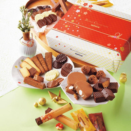 ROYCE' Chocolate - ROYCE' Collection "New Year" - Image shows a printed box in white and red with floral prints. Text on the box says ROYCE' Collection ROYCE'. Accents include wooden and ceramic plates filled with confections of varying colors and shapes. Other accessories seen are a white vase with flowers and some gold orbs and paper figurines. Background is a mix of white and green.