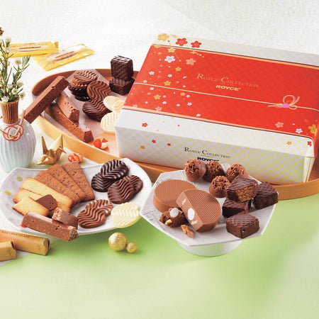ROYCE' Chocolate - ROYCE' Collection "New Year"  - Image shows a printed box in white and red with floral prints. Text on the box says ROYCE' Collection ROYCE'. Accents include wooden and ceramic plates filled with confections of varying colors and shapes. Other accessories seen are a white vase with flowers and some gold orbs and paper figurines. Background is a mix of white and green.