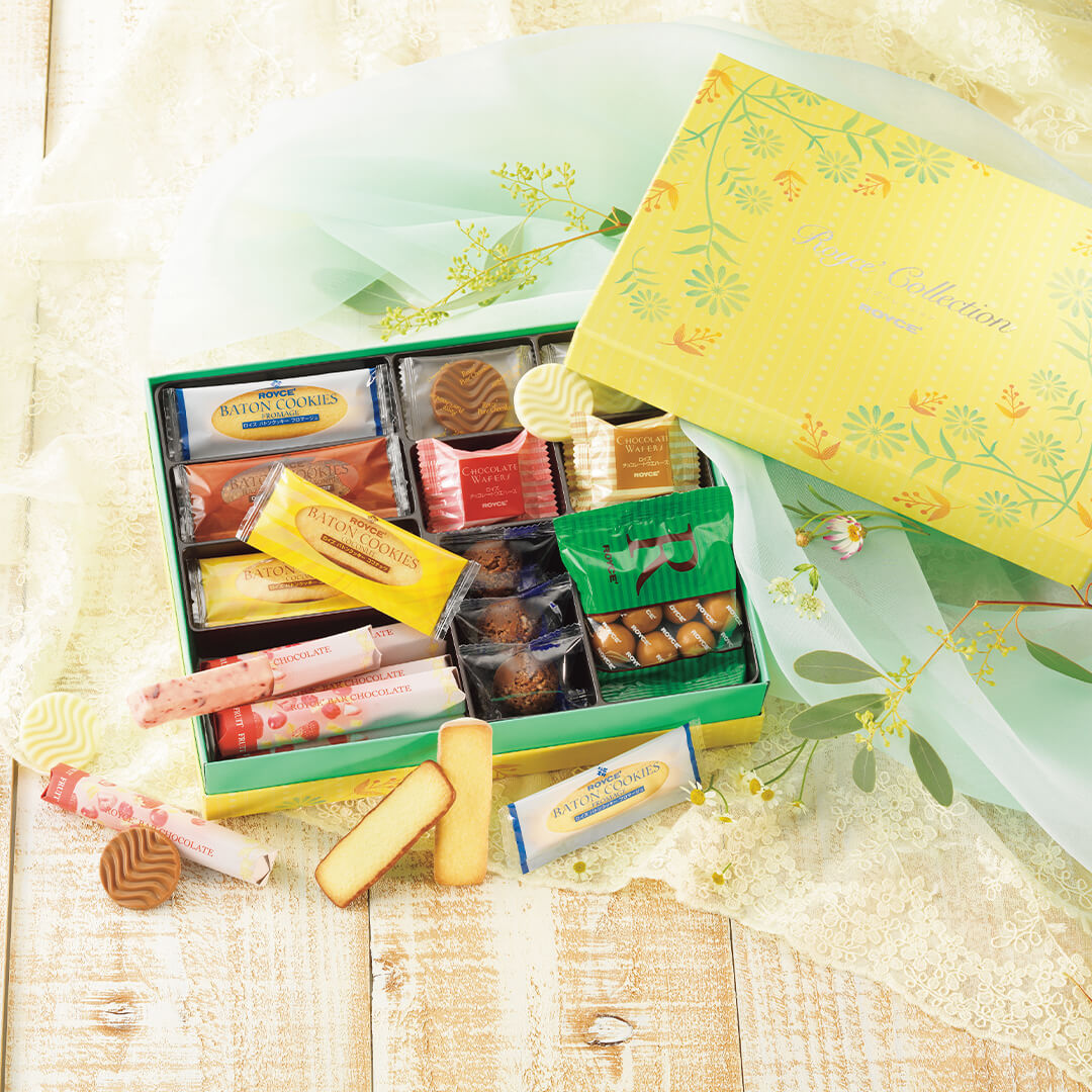 ROYCE' Chocolate - ROYCE' Collection "Sweet Yellow" - Image shows an open box with different confections inside that come in varying shapes, colors, and sizes. The box cover seen on the upper right portion is colored yellow with green and yellow floral prints. Text says ROYCE' Collection ROYCE'. Box below is in the colors green and yellow. Accents include a tablecloths in green and white and some green leaves and white flowers.