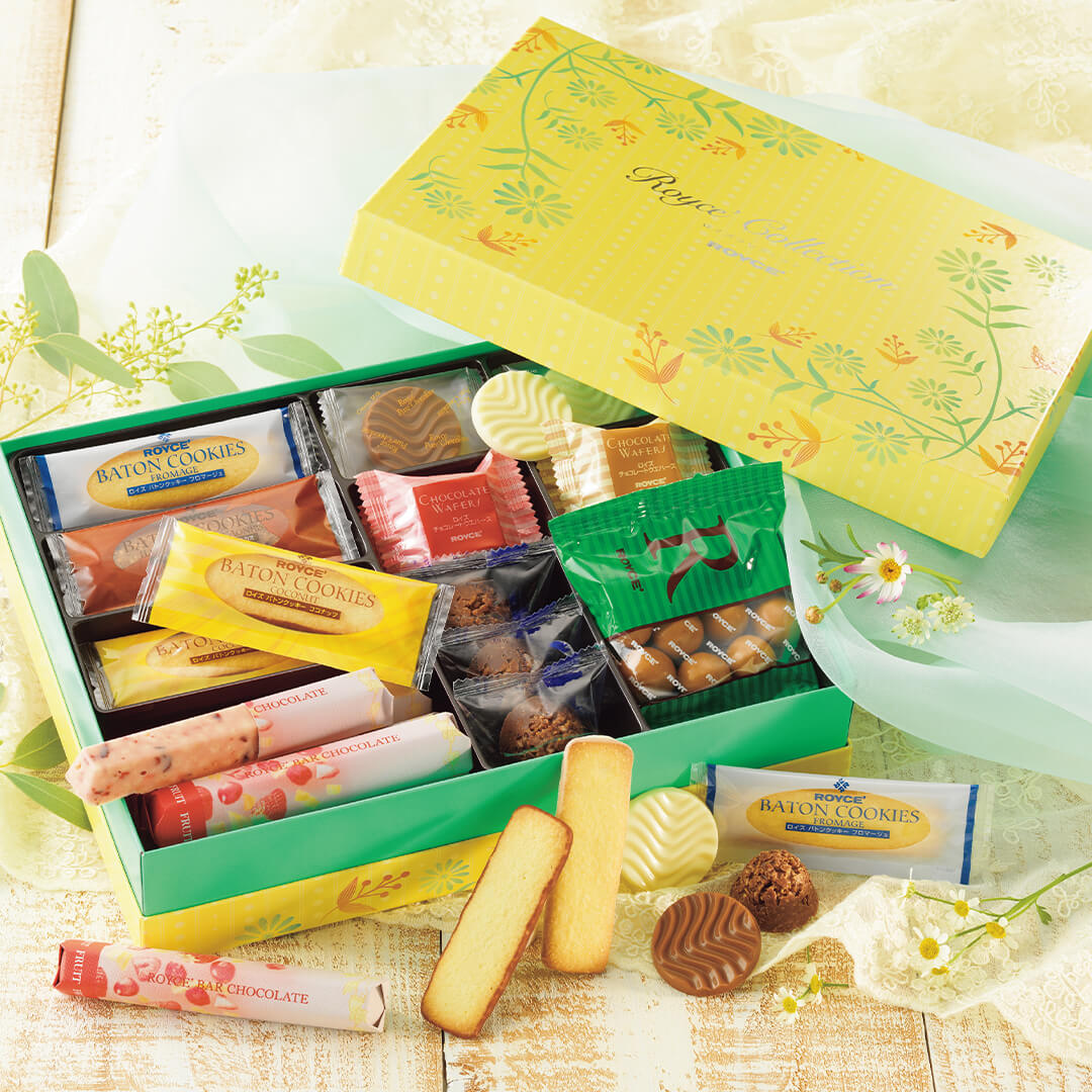 ROYCE' Chocolate - ROYCE' Collection "Sweet Yellow" - Image shows an open box with different confections inside that come in varying shapes, colors, and sizes. The box cover seen on the upper right portion is colored yellow with green and yellow floral prints. Text says ROYCE' Collection ROYCE'. Box below is in the colors green and yellow. Accents include a tablecloths in green and white and some green leaves and white flowers.