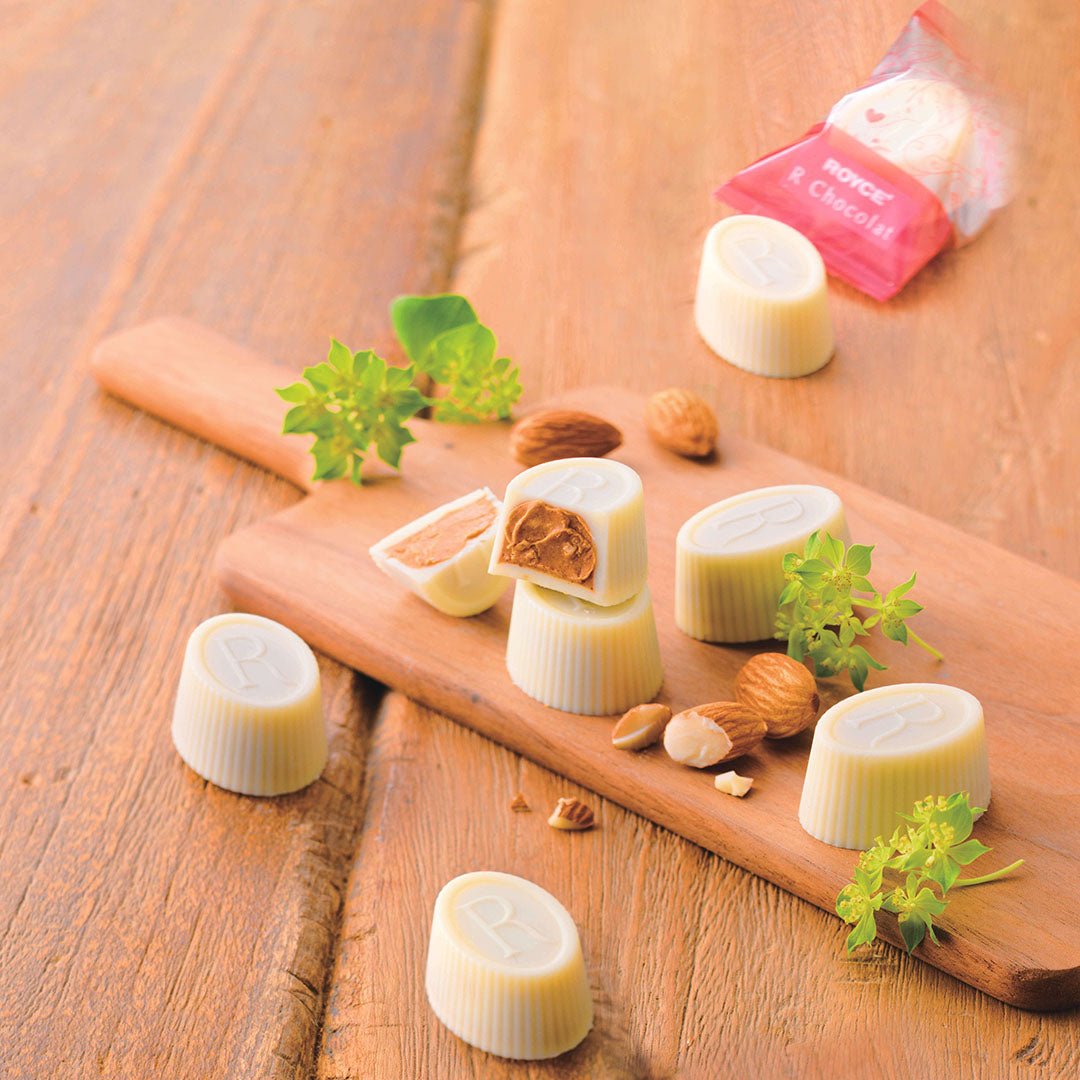 ROYCE' Chocolate - ROYCE' R Chocolat "White Gianduja" - Image shows white chocolate cups filled with a brown cream. Accents include a brown wooden chopping board and green leaves. Background and surface is brown wood.