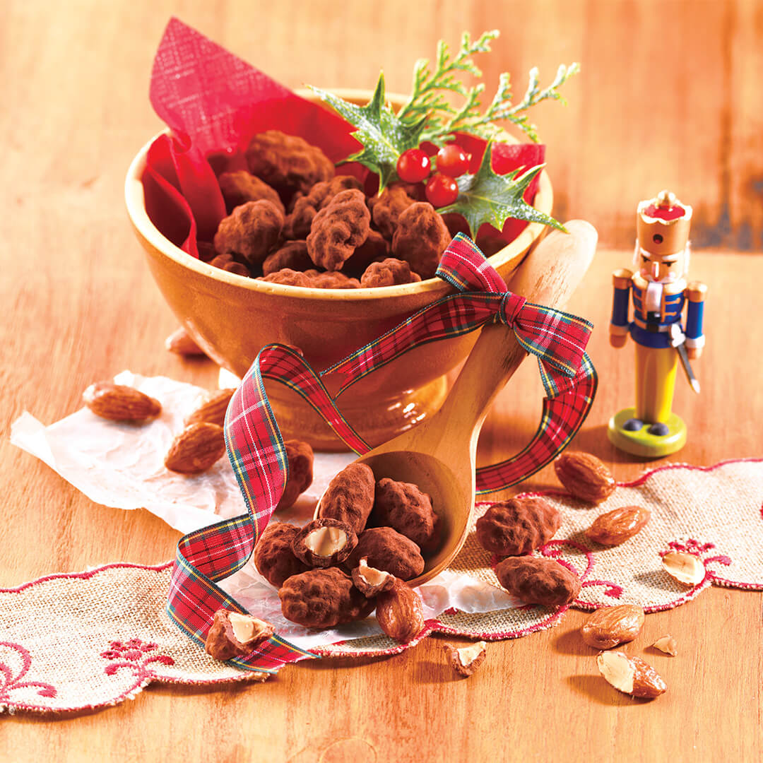 Roasted Almond Chocolate in a wooden bowl with cocoa-dusted, chocolate-covered almonds, a plaid-ribbon scoop, holly sprig, and toy nutcracker on an embroidered cloth.