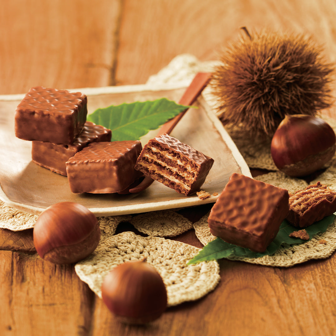 Image shows brown chocolate wafers on a brown plate and on a brown table with a crocheted placemat in light yellow. Accents include green leaves and chestnuts in brown. Background is in brown.