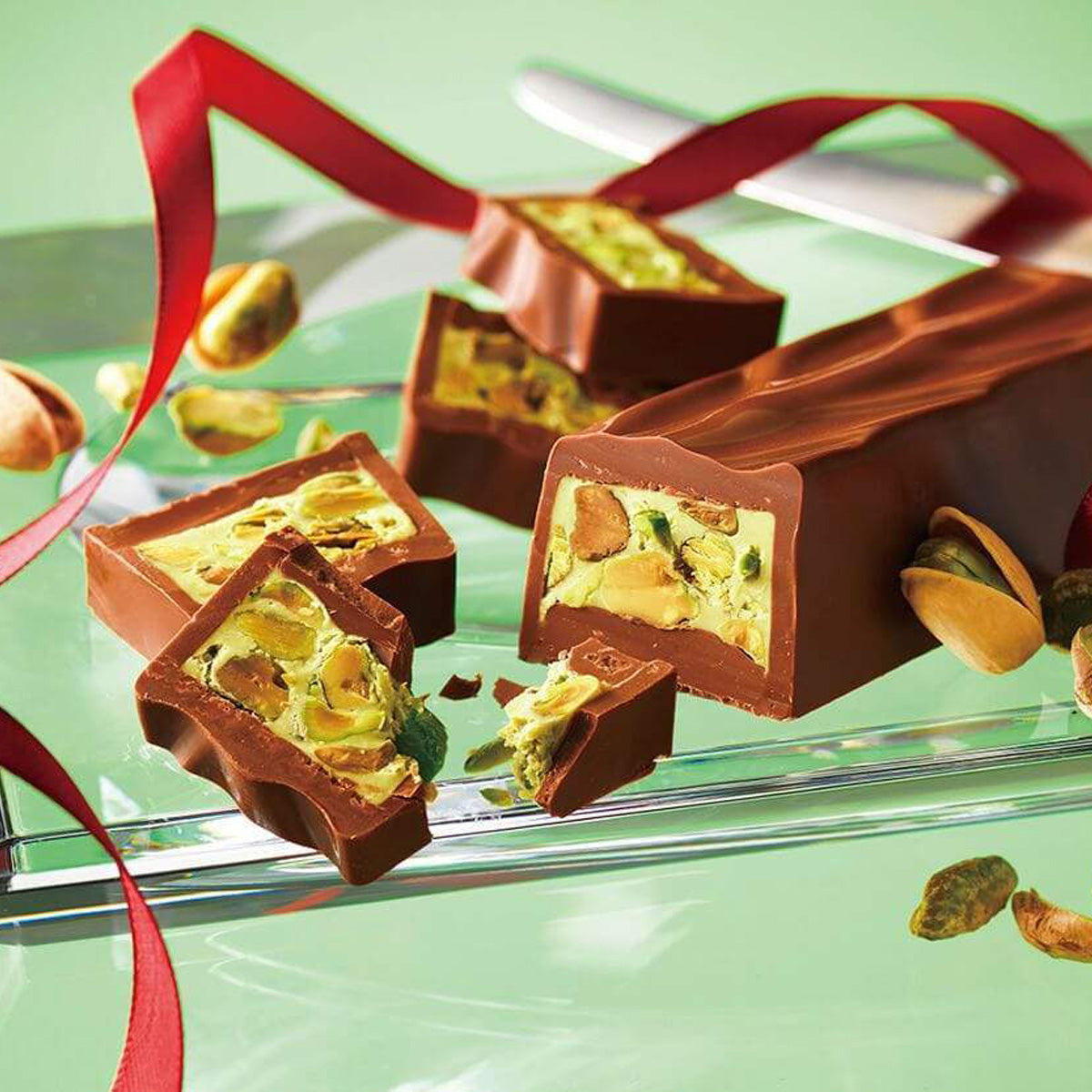 ROYCE' Chocolate - Pistache Chocolat - Image shows a brown chocolate loaf and slices filled with pistachios and green cream. Accents include a clear tray and red ribbon. Background is green.