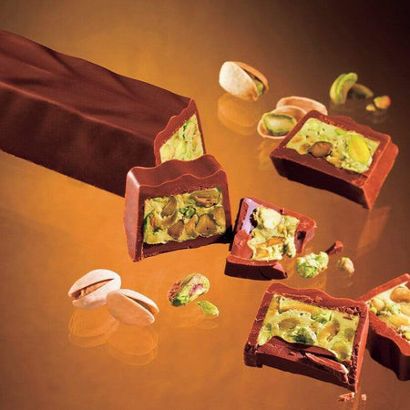 ROYCE' Chocolate - Pistache Chocolat - Image shows a brown chocolate loaf and slices filled with pistachios and green cream. Accents include loose pistachio nuts. Background is brown.