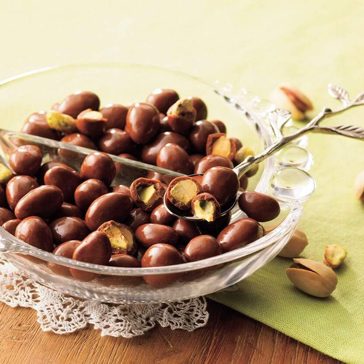 ROYCE' Chocolate - Pistachio Chocolate - Image shows a clear bowl filled with brown chocolate-coated pistachios. Accents include a silver spoon, white lace, and green tablecloth on wooden table.