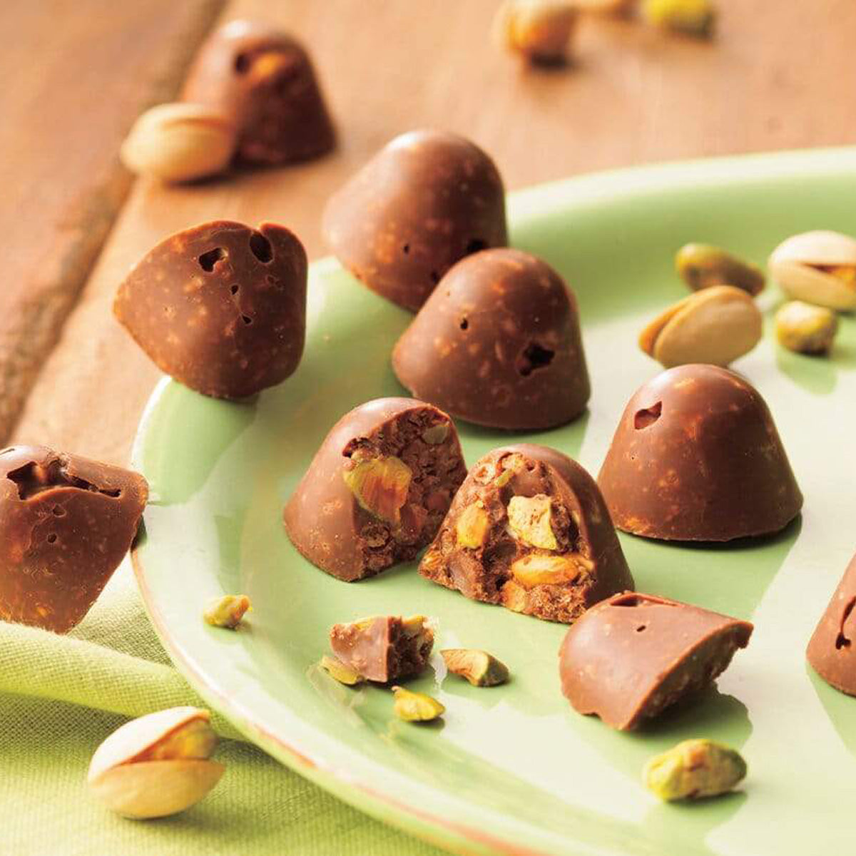 ROYCE' Chocolate - Pistachio Crunch Chocolate - Image shows a green plate filled with brown chocolate creations with pistachios. Accents include loose pistachios and a green tablecloth on wooden table.