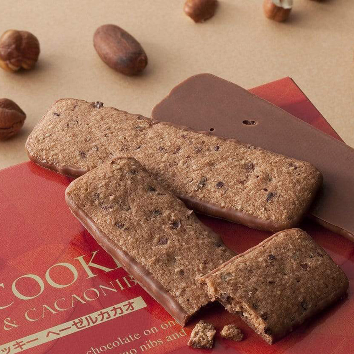 ROYCE' Chocolate - Baton Cookies "Hazel Cacao" - Image shows brown cookies with brown chocolate chips coated with brown chocolate on top of a red box with letters. Accents include nuts in various shapes. Background is brown.