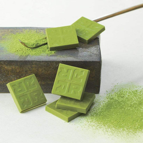 Matcha Chocolate (32 Pcs) features green chocolate squares with a dusting of matcha powder, arranged on a metal block beside a bamboo tea whisk tip.